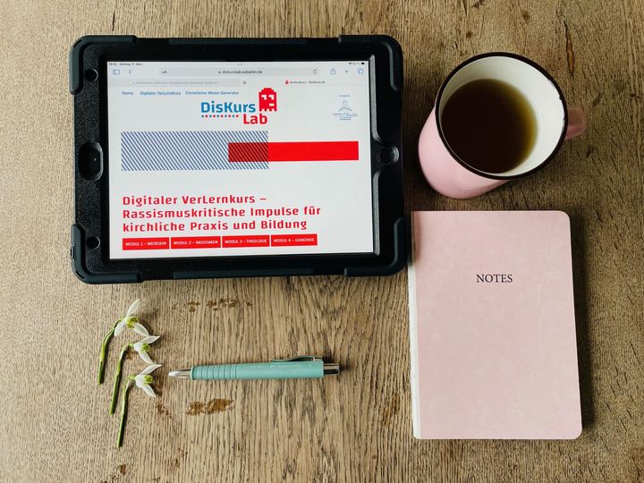 Auf einem Tisch liegen ein Stift, ein Notizbuch, eine Tasse und Tablet mit Hinweis auf Rassismus-Verlernkurs vom DiskursLab.