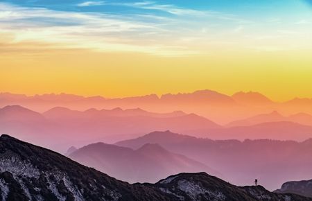 Bergmassiv getaucht in regenbogenfarbendes Licht, vorne steht klein auf einem Bergkamm ein Mensch