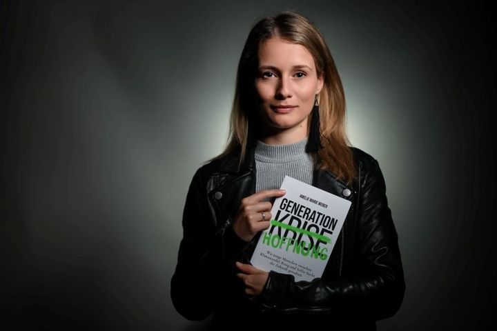Eine Frau mit langen Haaren hält ein Buch in der Hand und schaut in die Kamera.