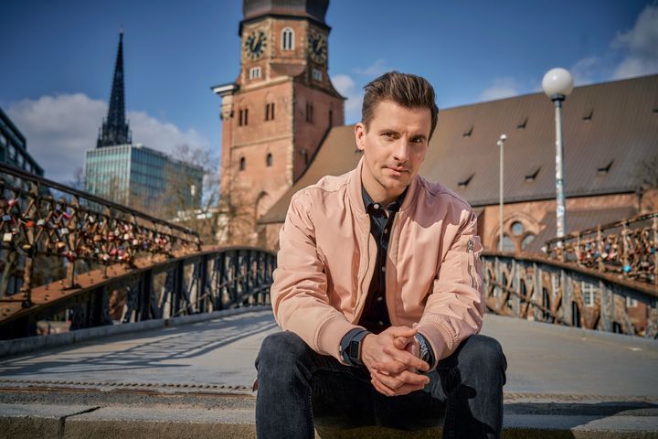Ein Mann mit rosefarbener Jacke sitzt im Freien, im Hintergrund eine Brücke, blauer Himmel und Gebäude.