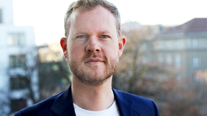 Ein Mann mit Jacket und weißem Kragen schaut in die Kamera. Im Hintergund sind unscharf Häuser zu erkennen.