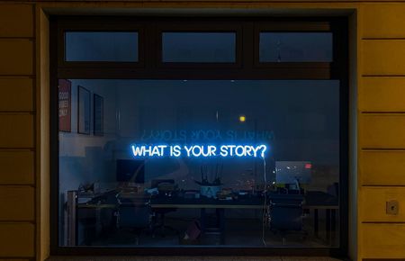 Dunkle Fensterscheibe auf der ein blauer Neonschriftzug "What is your story?"