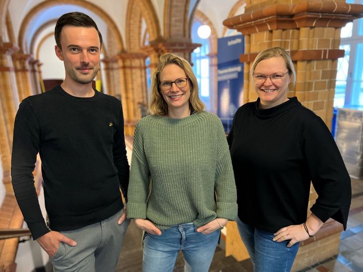 Drei Personen stehen in einem Treppenhaus und schauen freundlich in die Kamera.