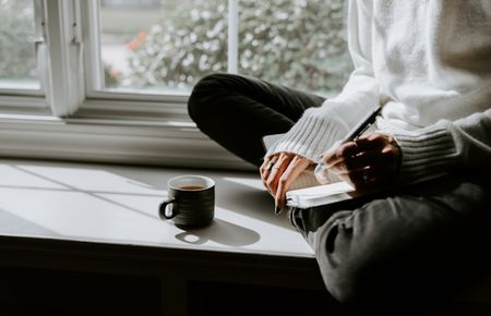 Auf der Fensterbank sitzt bei Sonnenlicht eine Frau im weißen Pulli mit Notizbuch, Stift, Tasse - davon ist ein Ausschnitt zu sehen