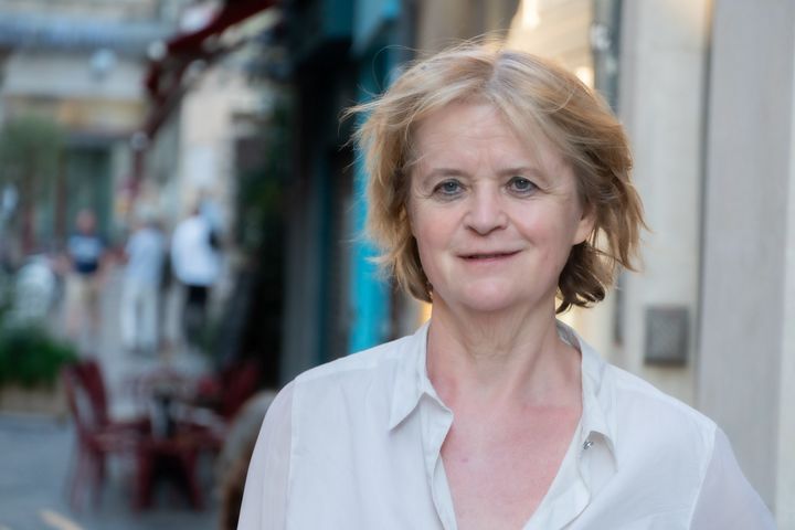 Eine Frau mit weißer Bluse schaut freundlich in die Kamera. Im Hintergund unscharf eine Straße mit Gebäuden und Menschen.