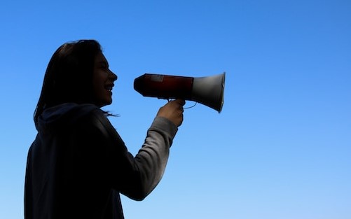 vor strahlend blauem Himmel ist eine weibliche Silhoutte sichtbar, die ein Megaphone hält und spricht