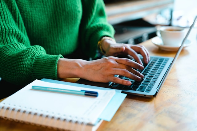 kit/ unsplash Ausschnitt einer Frau im grünen Strickpulli, die ihre Hände am Laptop hat, daneben ein Notizblock mit Stift