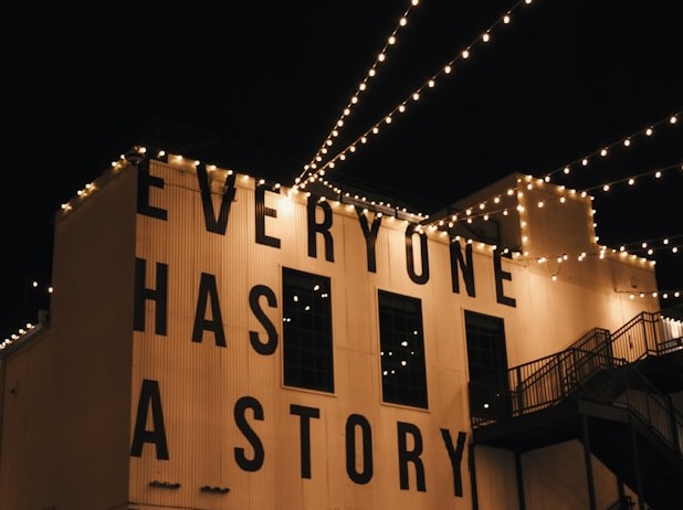 Liliy Miller/ unsplash Häuserwand bei Nacht mit Lichterketten und der Aufschrift: Everyone has a story